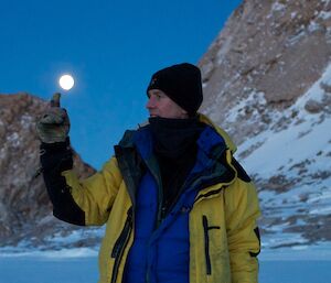 Expeditioner with fingered glove pointing upwards seeming to balance the rising moon