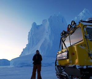 A female expeditioner on the sea ice, standing near a yellow Hägglunds with an iceberg in the background.