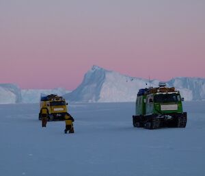 In the foreground there are two Hägglunds and two people with icebergs behind and a pastel pink sky in the morning