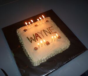 A roughly square cake decorated with 12 candles and Wayne’s name placed on a silver papered covered board