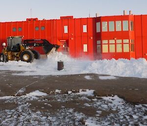 An IT Loader moving snow with the red main living and sleeping quarters behind