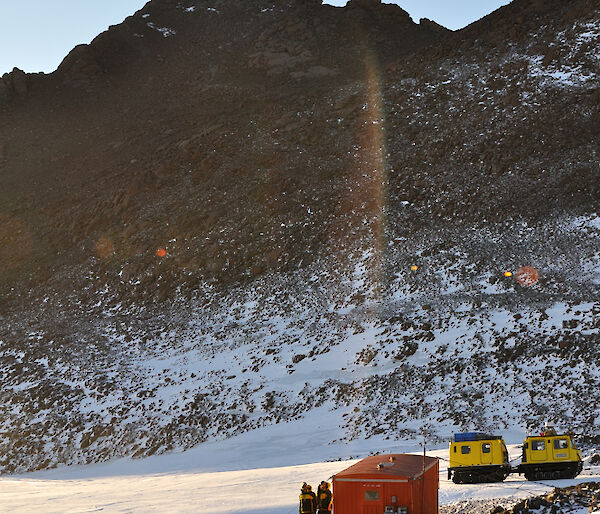 A small orange field hut with 2 expeditioners on the verandah and a Yellow H agglunds behind the hut and the summit of Mt Henderson looming above