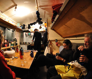 The inside of the Macey Island field hut with 3 expeditioners sitting on the beds, an expeditioner standing near the stove and glasses on the table