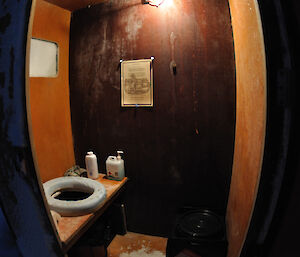 A Fish Eye lens photo of the Macey toilet showing a toilet seat, with talcum powder and hand wash on the bench and a waste container for urine on the floor