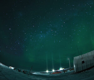 Three vertical pillars of light shine from outside station lights in the night sky filled with aurora