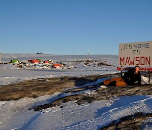 A photo of an expeditioner at the Mawson welcome sign with the station in the middle distance