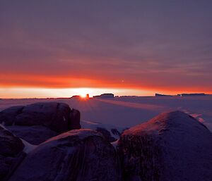 The sky is red with the sun on the horizon and beautiful reflections on the sea-ice and rocks on an island in the foreground