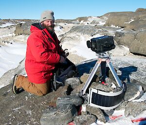 An expeditioner beside one of the penguin nest cameras showing solar panel, tripod and pelican case enclosing the camera with the shutter open