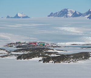 A photo showing the coloured buildings of Mawson in the middle of the photo with the ice plateau and the mountains of the David Range and spectacular twin peaks of Mt Hordern