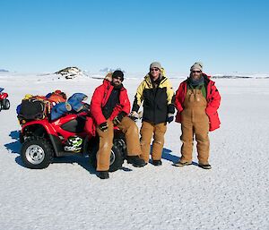 Three expeditioners standing beside their quads on the sea-ice with the Antarctic plateau behind on a sunny day