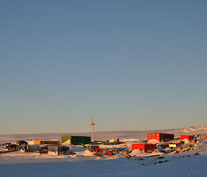 A photo of mawson on a clear blue sky with the Antarctic plateau behind and sea-ice in front of teh various coloured buildings of the Station