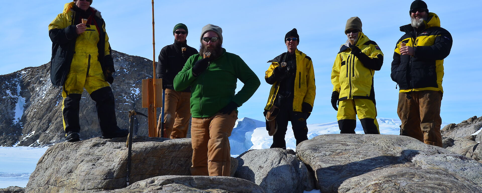 A group of six expeditioners surrounding Mawson’s Proclamation Plaque with ice creams in their hands