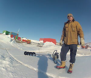 An expeditioner standing on the sea ice with a jiffy drill at his feet and the buildings of Mawson Station in the background