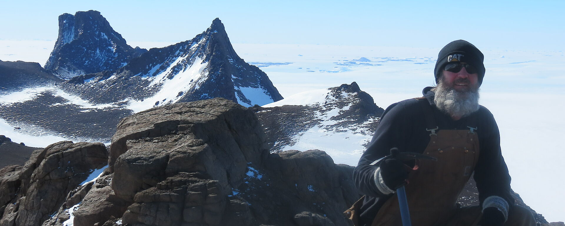 An expeditioner on a rocky platform with the tooth like summit of Fang Peak and another summit behind him