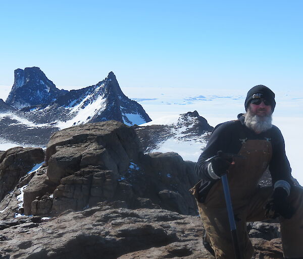 An expeditioner on a rocky platform with the tooth like summit of Fang Peak and another summit behind him