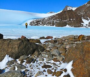 An expeditioner walking on a frozen lake bounded by an ice slope, mountain and different colored rocks in a moraine