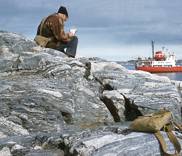 A scientist sits on a rock overlooking a bay writing in his field notebook.