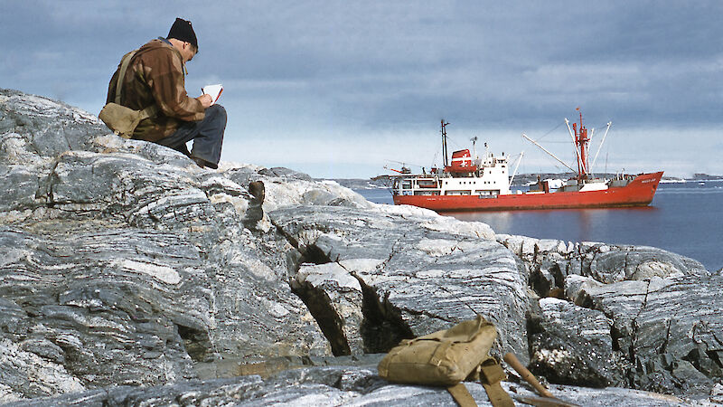 A scientist sits on a rock overlooking a bay writing in his field notebook.