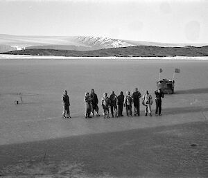 Black and white image of men lined up on the fast ice at Mawson in 1954 farewelling ship. Weasel and coastline in background.
