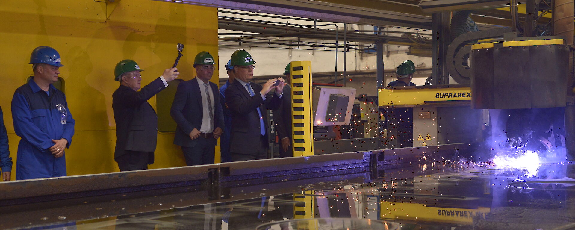 People standing around vat of water with machine cutting steel and throwing flames off