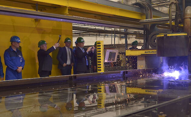 People standing around vat of water with machine cutting steel and throwing flames off