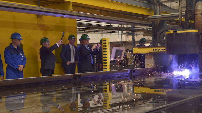 People standing around vat of water with machine cutting steel and throwing flames off