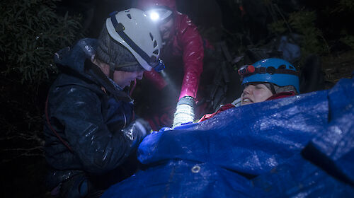 Mock patient wrapped in blanket being tended by two people.