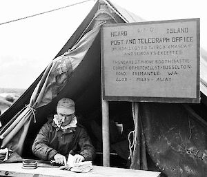 Black and white photo of a man sitting in front of a tent with a post office sign above his head.