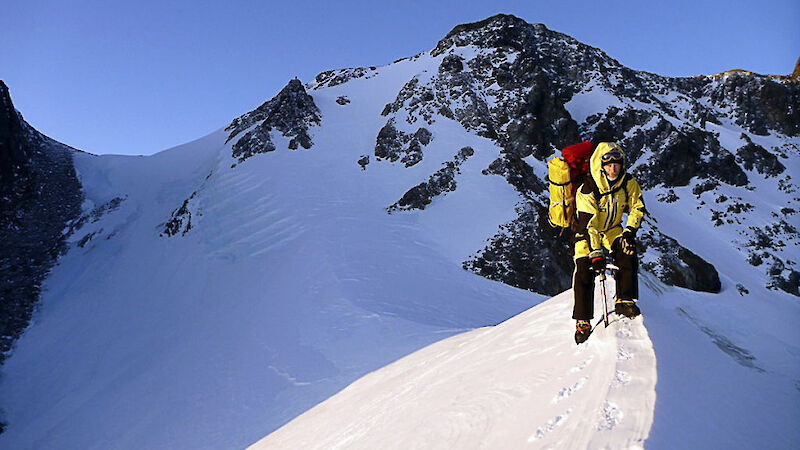 Expeditioner walking along a snow line with a mountain behind.