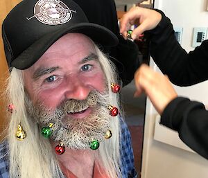 Man with long white hair and black cap having Christmas decorations put into his beard.