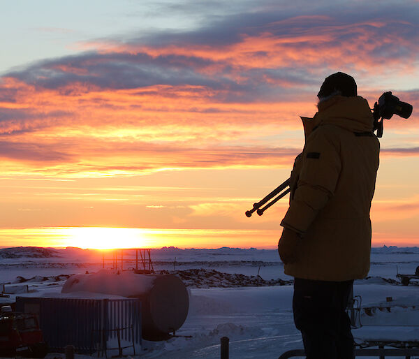 Expeditioner with camera tripod over shoulder walks in front of rising sun