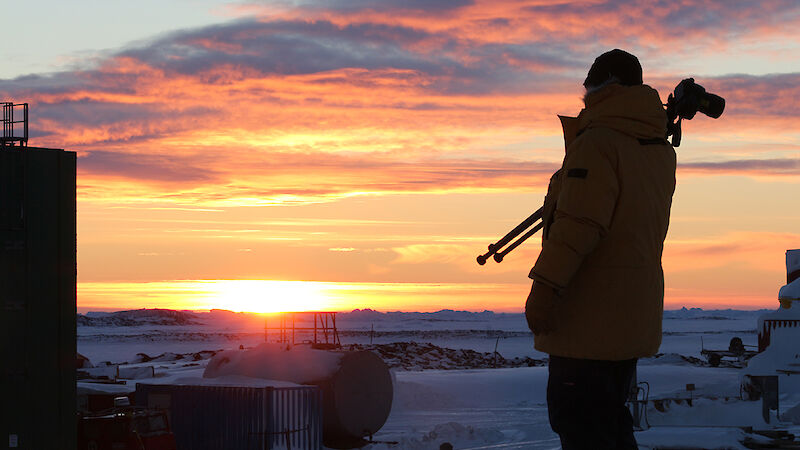 Expeditioner with camera tripod over shoulder walks in front of rising sun