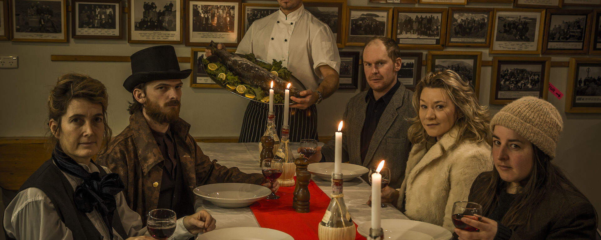 Expeditioners in historic clothes sit around a table with serious faces while a chef presents a fish on a platter