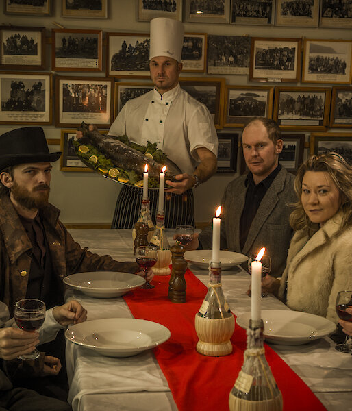 Expeditioners in historic clothes sit around a table with serious faces while a chef presents a fish on a platter