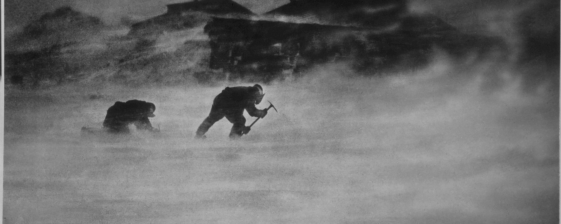 Historic image of expeditioners leaning into a ferocious wind whilst chipping ice to melt for water