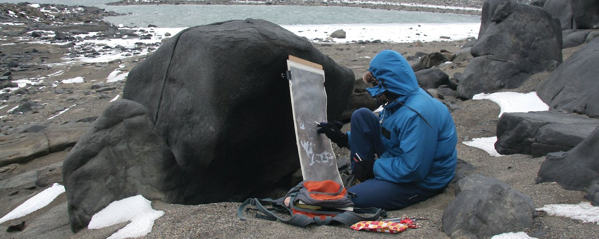 A person hunches on the ground painting with the canvas resting against a rock.