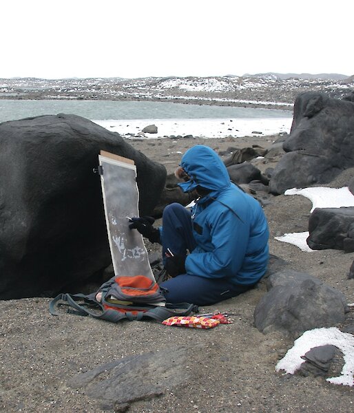 A person hunches on the ground painting with the canvas resting against a rock.