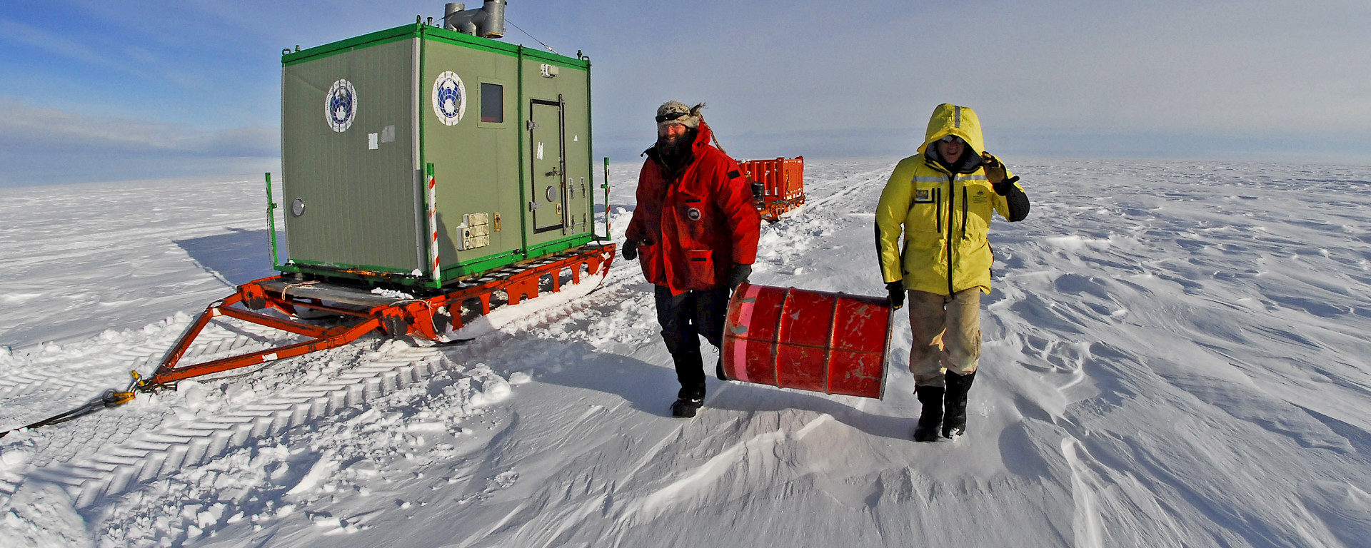 Two mean carrying drums across the ice with a green trailer behind them