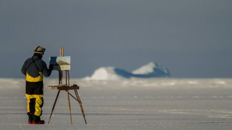 JOhn Kelly wearing protective Antarctica gear, paints with an easel set up on the ice,