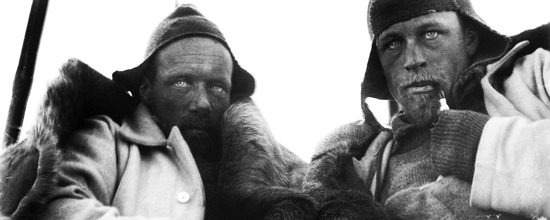 Two men wearing woollen beanies and hunching to keep warm stare at the camera with weathered faces.