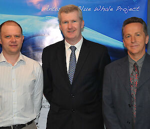 Three men stand smiling at the camera with a banner of a blue whale behind them