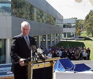 Governor-General at lectern with rows of seated staff on lawn behind.