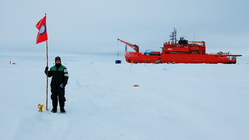 Alex standing on the sea ice and the Aurora Australis behind, during the Sea Ice Physics and Ecosystem eXperiment voyage in 2007.