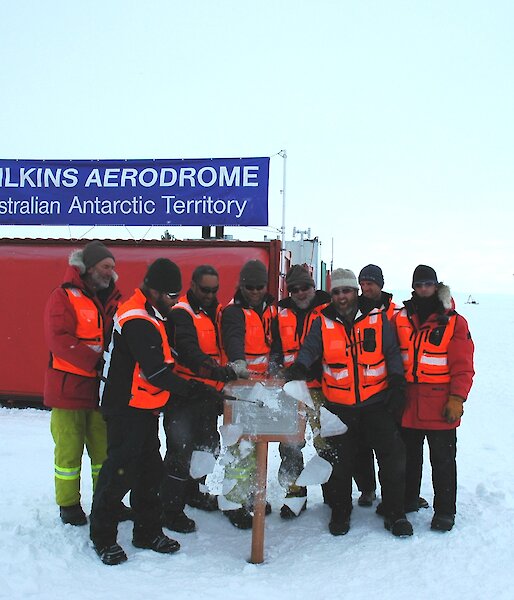 Eight expeditioners unveiling a plaque at Wilkins Runway as they cracked the ice encasing it.