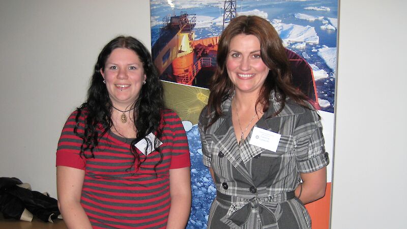 Two female teachers stand in front of a poster of the research vessel, Aurora Australis.