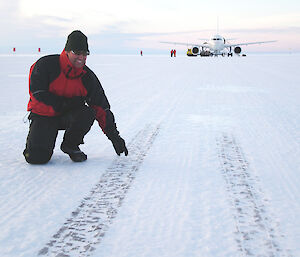 Person wearing high vis pointing at tyre marks on an ice runway with a plane in the background.