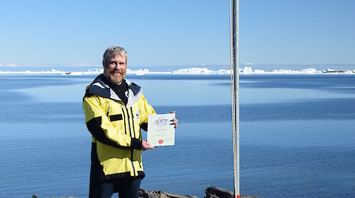 Man wearing a yellow jacket standing under a flag holding a certificate.