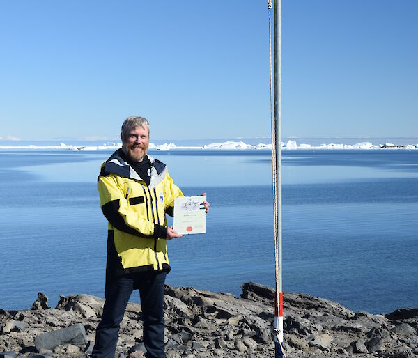 Man wearing a yellow jacket standing under a flag holding a certificate.