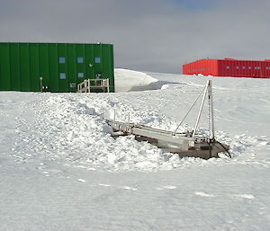 A raised metal walkway in snow from a green building at Casey station