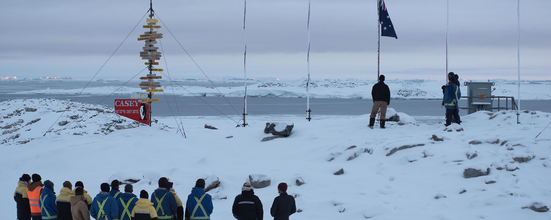 Casey research station expeditioners gather in front of the flag poles for Anzac Day service